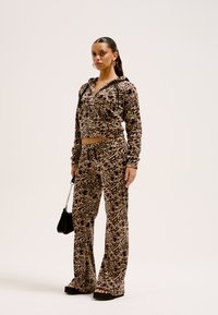Afskåret, lynlås-hoodie og vide bukser i brunt leopardprint-stof, der har en blød tekstur og sidelommer. Inkluderer en lille sort håndvæg.