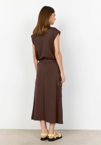 Brown, short-sleeve dress with a relaxed fit, featuring two side pockets and a drawstring waist. Worn with yellow strappy sandals.