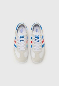 Sneakers com parte superior em couro branco, pormenores em camurça cinza, riscas sintéticas em azul e vermelho, atacadores brancos e palmilha almofadada.
