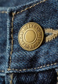 Țesătură din denim, de culoare albastru închis cu cusături vizibile, având un buton metalic cu inscripția "WEEKDAY." Prim-planul arată textura și detaliile.