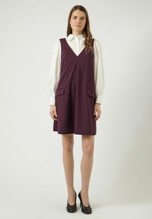 Pitkätukkainen nainen, joka pukeutunut valkoiseen puseroon, jossa on pulleat hihat, purppuraan hihattomaan mekkoon ja mustiin korkokenkiin, seisoo yksiväristä taustaa vasten.