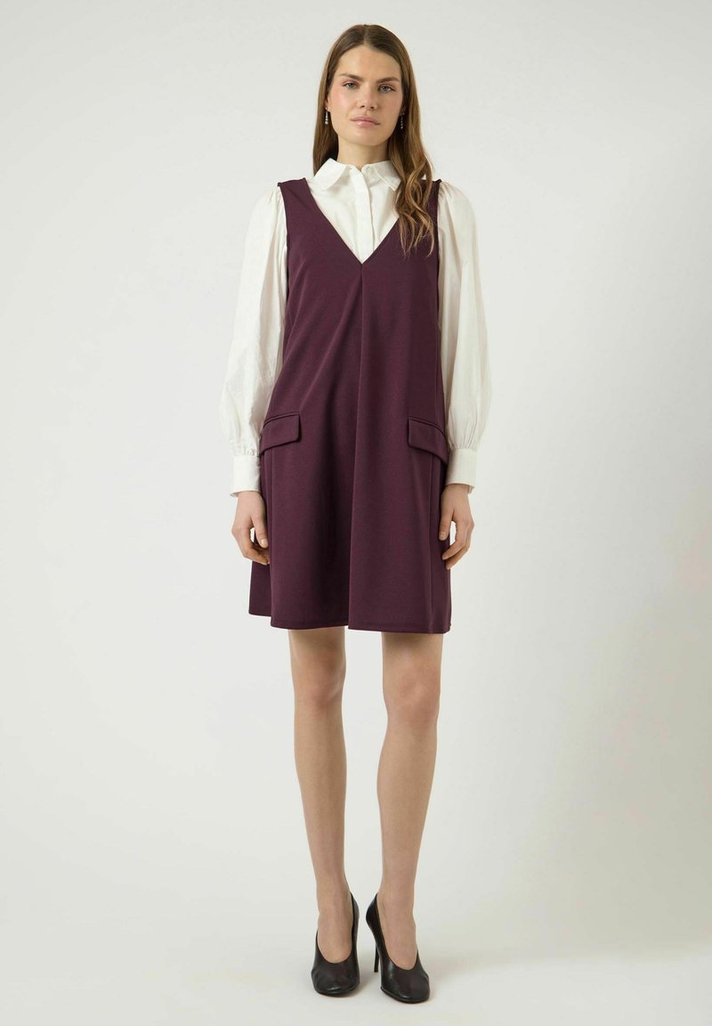 Mujer con cabello largo y castaño, vestida con una blusa blanca de mangas abullonadas bajo un vestido morado sin mangas y tacones altos negros, posando frente a un fondo liso.