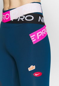 Granatowe legginsy sportowe mają szeroki pas w różowe, czarne i białe logo "PRO", a także białe i czerwone akcenty z logo Nike.