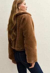 Brązowa kurtka z sztucznego futra o krótkim kroju, szerokich rękawach i miękkiej fakturze, w zestawieniu z ciemnymi jeansami.