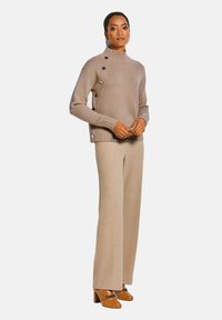 Beige Strickpullover mit hohem Kragen und Knopfakzenten an der Seite, kombiniert mit beigen, weit geschnittenen Hosen und braunen Absatzschuhen.