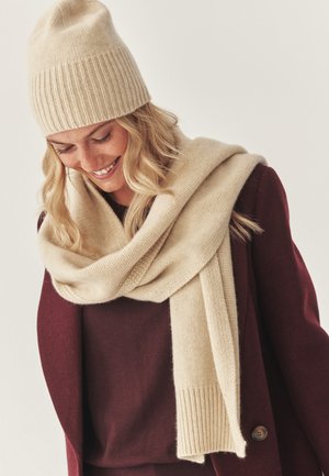 Cream-colored knitted beanie with ribbed pattern and matching scarf draped over a maroon sweater. Soft texture, warm appearance, and simple design.