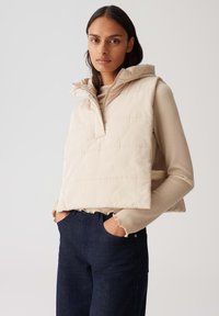 Beige gesteppte Weste mit hohem Kragen und halbseitigem Reißverschluss, über einem hellbraunen gerippten Oberteil getragen, kombiniert mit dunkelblauen Jeans.