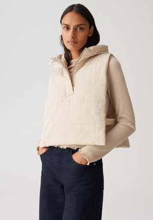 Gilet matelassé beige avec un col haut et une fermeture éclair mi-hauteur, superposé à un haut côtelé beige clair, associé à un jeans bleu foncé.