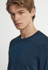 Marinblå rundhalsad tröja med fin struktur; figursydd passform framhäver axlarna; subtil lyster reflekterar ljus; minimalistisk design.