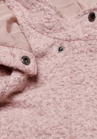 Tessuto testurizzato rosa di una giacca con un esterno soffice, dotata di una fodera interna liscia, bottoni a pressione in metallo e dettagli di cucitura sottili.