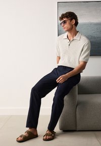 Hellgraues Poloshirt mit kurzen Ärmeln, marineblaue Hose, braune Sandalen. Das Modell hat lockiges Haar und trägt eine Sonnenbrille. Modernes, minimalistisches Ambiente.