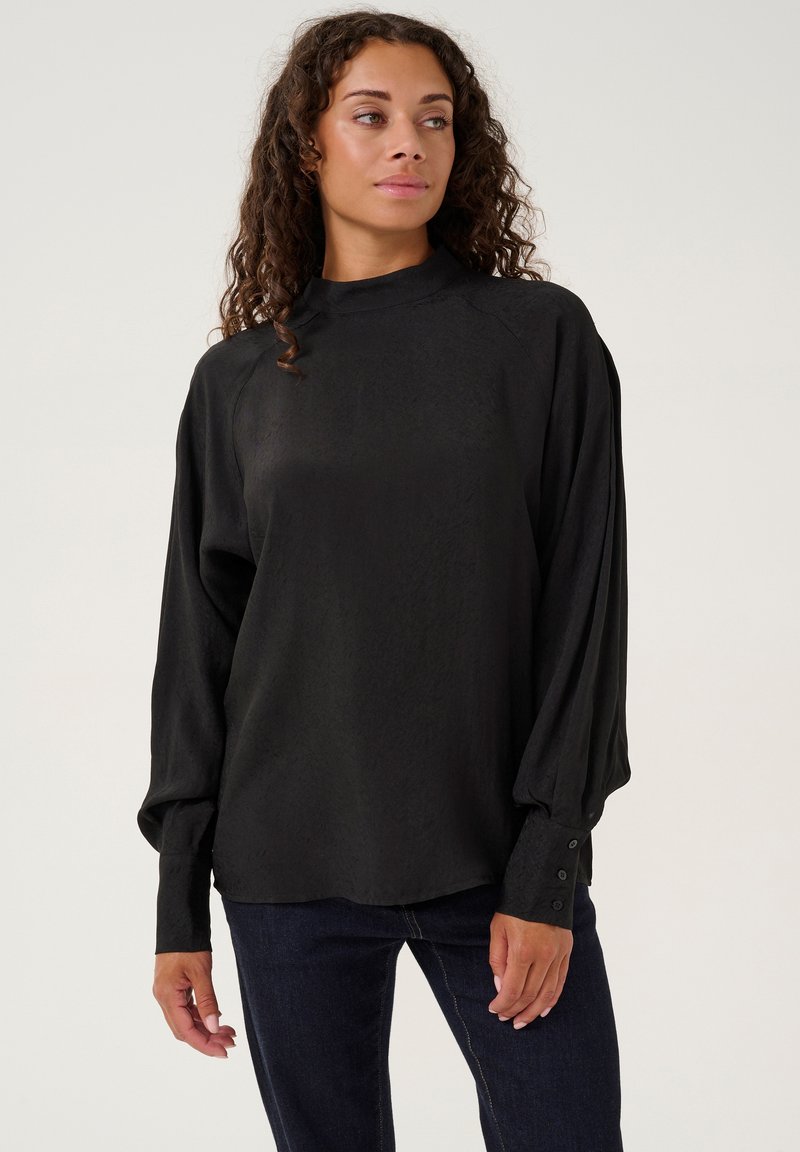 Blusa nera con collo alto, maniche lunghe a sbuffo e dettagli a bottoni sui polsini. Realizzata in un tessuto liscio con una sottile texture.