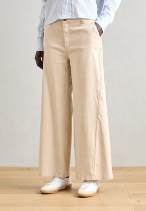 Pantalons larges beige en tissu doux, dotés d'une fermeture éclair à l'avant, de poches latérales et d'un ourlet net, associés à des baskets blanches.