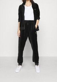 Osoba oblečená v černém sametovém teplákovém obleku s kardiganem na zip a kalhotami se šňůrkou, bílém tílku a bílých teniskách, stojící proti jednoduchému pozadí.