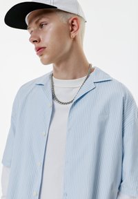 Jeune homme aux cheveux blond clair portant une casquette blanche, un collier en chaîne argenté, une chemise rayée bleu clair par-dessus un haut à manches longues blanc, regardant à gauche.
