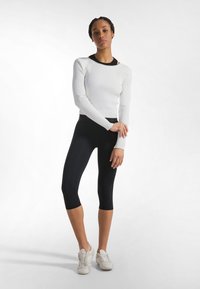 Weißes, geripptes Langarmshirt mit schwarzem Ausschnitt, kombiniert mit enganliegenden schwarzen Capri-Leggings und weißen Sportschuhen. Gleichmäßige Textur überall.