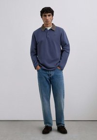 Men's long-sleeve polo shirt in blue with a brown collar, paired with light blue jeans and dark brown shoes. Simple design and relaxed fit.