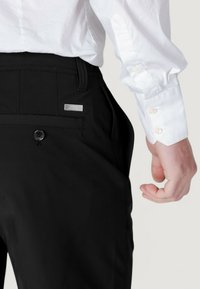 Pantalones negros con una textura suave, que incluyen un bolsillo lateral, un cierre de botón y una pequeña etiqueta metálica en la cintura.