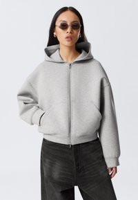 Grå zip-up hoodie med en stor huva, oversize passform och framfickor. Kombinerad med mörka denimbyxor, visad mot en neutral bakgrund.