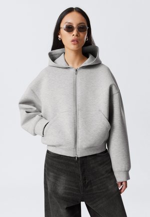 Sudadera gris con cremallera, capucha grande, corte oversize y bolsillos frontales. Combinada con pantalones de mezclilla oscuros, presentada sobre un fondo neutro.