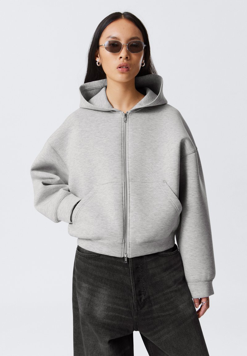 Grå zip-up hoodie med en stor huva, oversize passform och framfickor. Kombinerad med mörka denimbyxor, visad mot en neutral bakgrund.