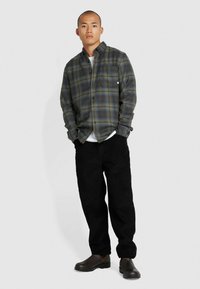 Geruite flanellen shirt in groene, blauwe en zwarte tinten, met knopen en een borstzak, gecombineerd met zwarte corduroybroek en donkere schoenen.