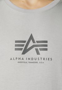 Pilka medvilninė marškinėliai su tamsiai pilku logotipu, kuriame stilizuotas „A“ simbolis ir horizontalių juostelių. Tekstas skaito: "ALPHA INDUSTRIES, KNOXVILLE, TENNESSEE, JAV."