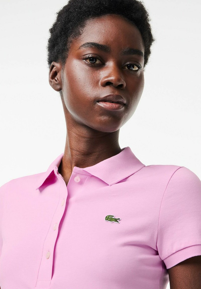 Frau mit kurzen lockigen Haaren, die ein hellrosa Polohemd mit einem grünen Krokodil-Logo auf der linken Brust trägt, und leicht nach oben schaut.