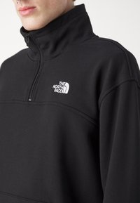 Sudadera de forro polar negra con cremallera de cuarto y cuello alto, que presenta un pequeño logo blanco de The North Face en el lado izquierdo. Textura suave, diseño minimalista.