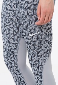 Šedé sportovní legíny s geometrickým černým a šedým vzorem, se světle šedými akcenty a bílým logem Nike na boku.