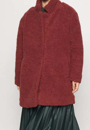 Abrigo rojo, esponjoso y sobredimensionado con alto cuello, tejido texturizado, cierre delantero y mangas largas, combinado con una falda plisada verde oscuro.