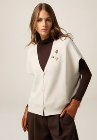 Creme korte mouwen cardigan met een diepe V-hals, knoopsluiting en decoratieve broches. Gecombineerd met een bruine coltrui en bruine broek.