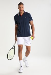 Marineblauw polo-shirt met rode en witte accenten, gecombineerd met witte shorts en witte sportschoenen. Houdt een tennisracket en bal vast.