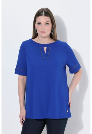 Frau steht vor einer weißen Wand, trägt ein blaues kurzärmliges Oberteil mit schwarzem Besatz und schwarze Hosen, blickt nach vorne mit neutralem Gesichtsausdruck.