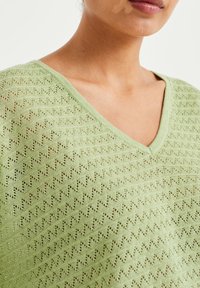 Vihreä neulepaita, jossa on V-pääntie ja teksturoitu sahalaitamalli sekä hienovaraisia avoimia silmukoita hengittävyyden lisäämiseksi.