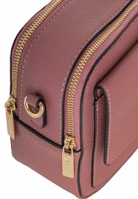 Skórzana torba w miękkim mauve z teksturowanym wykończeniem. Posiada złote suwaki, czarne akcenty oraz boczną kieszeń z metalowym detalem w formie pierścienia.