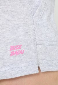 Šedá bavlněná látka má boční šev a malou růžovou logo „BTDI BADU“ blízko lemu, které zdůrazňuje jednoduchý design.