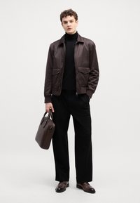 Veste bombers marron, pull noir à col roulé, pantalon noir et chaussures en cuir marron. Le mannequin tient un sac à main marron assorti avec une texture lisse.