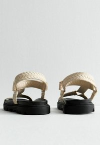 Sandali beige intrecciati con una parte superiore testurizzata, cinturini regolabili e una suola nera piatta. Presentano dettagli cuciti e una soletta imbottita.