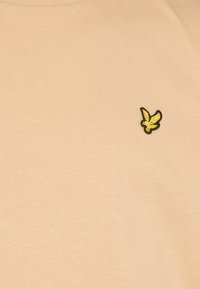 Beige, rövid ujjú póló sima anyagból, bal mellen egy kisebb fekete és sárga hímzett madárlogo található.