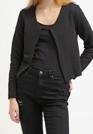 Femme portant un cardigan noir texturé à ouverture devant, sur un haut noir uni et un jean noir taille haute légèrement délavé.