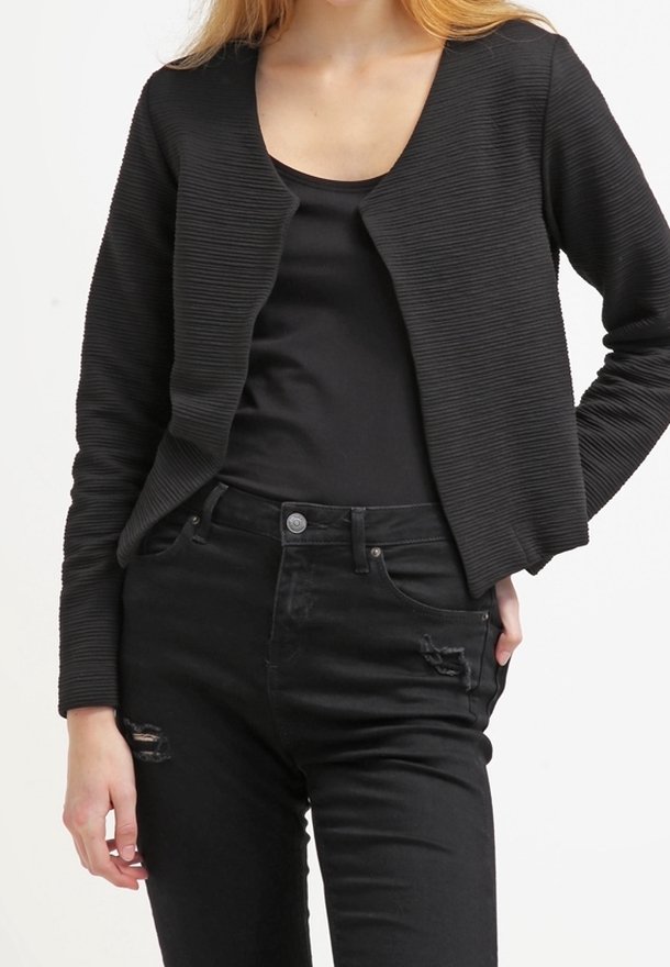 Femme portant un cardigan noir texturé à ouverture devant, sur un haut noir uni et un jean noir taille haute légèrement délavé.