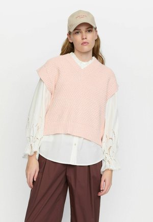 Gilet rose en tricot avec un col en V, des manches courtes et un ourlet côtelé, superposé sur une blouse blanche avec des détails en dentelle, associé à un pantalon marron.