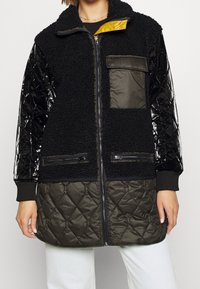 Svart fleecejacka med blanka svarta quiltade ärmar och khakifärgad nederdel. Har dragkedjefickor och ett gult foder. Dragkedjestängning.