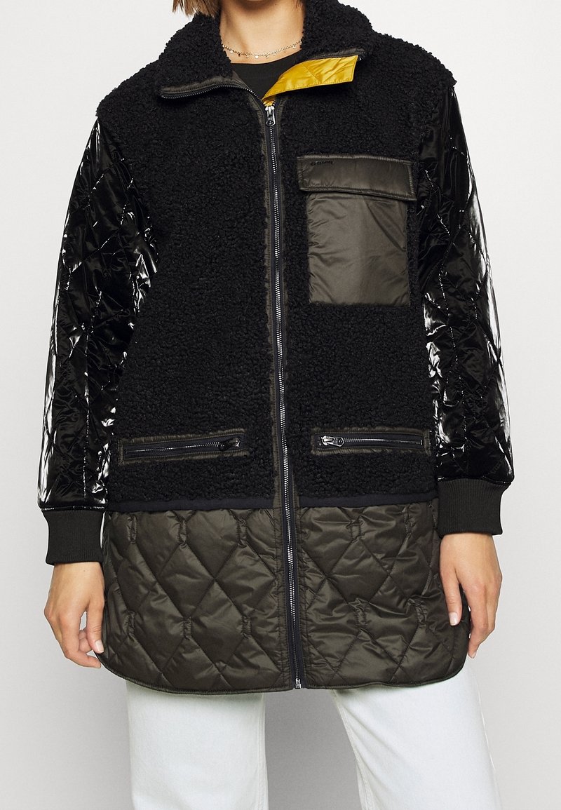 Svart fleecejacka med blanka svarta quiltade ärmar och khakifärgad nederdel. Har dragkedjefickor och ett gult foder. Dragkedjestängning.