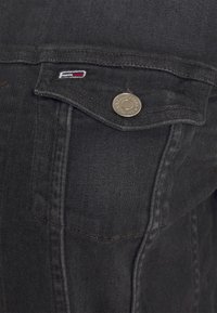 Czarna kurtka dżinsowa z małym haftowanym logo i srebrnymi zatrzaskami. Posiada kieszeń na piersi i widoczne przeszycia.