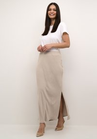 Rok maxi beige dengan belahan samping, terbuat dari kain bertekstur. Dipasangkan dengan kaus putih yang pas dan sepatu hak tinggi beige.