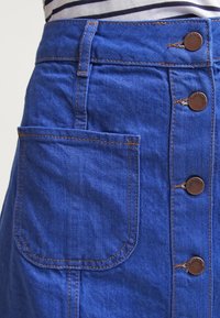 Modrá džínová sukně s přední kapsou, čtyřmi viditelnými měděnými knoflíky a oranžovým šitím, nošena s pruhovaným topem.