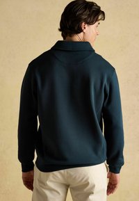 Vīrietis, ģērbies tumši zaļā garām piedurknēm svīterī ar apkakli, pagriežoties prom, valkā gaiši pelēkus bikses uz viendabīga pelēkā fona.