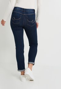 Jeans de mezclilla oscura con un corte ajustado, que presentan puños enrollados y costuras decorativas en los bolsillos traseros. Combinados con zapatos blancos sin cordones.